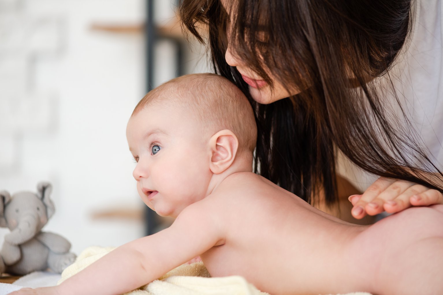Eine Frau mit langen braunen Haaren beugt sich sanft &uuml;ber ein nacktes Baby, das auf einem weichen gelben Handtuch liegt, und st&uuml;tzt es, w&auml;hrend es mit gro&szlig;en blauen Augen nach vorne schaut. Die Szene ist hell und zart.