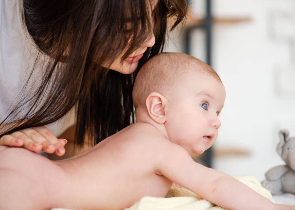 Eine Frau mit langen braunen Haaren beugt sich sanft &uuml;ber ein nacktes Baby, das auf einem weichen gelben Handtuch liegt, und st&uuml;tzt es, w&auml;hrend es mit gro&szlig;en blauen Augen nach vorne schaut. Die Szene ist hell und zart.
