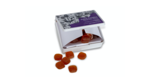 Ein wei&szlig;er Kunststoffbeh&auml;lter mit lila Etikett, der teilweise ge&ouml;ffnet ist und orangefarbene, rautenf&ouml;rmige Bonbons zeigt, wobei mehrere Bonbons vor einem wei&szlig;en Hintergrund versch&uuml;ttet sind.