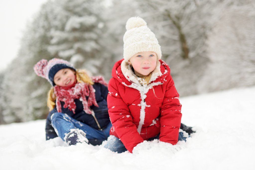 Zwei Kinder in warmer Winterkleidung sitzen im Schnee und spielen. Das Kind im Vordergrund tr&auml;gt einen roten Mantel und eine wei&szlig;e M&uuml;tze, w&auml;hrend das Kind im Hintergrund eine gemusterte M&uuml;tze, einen blauen Mantel und einen roten Schal tr&auml;gt. Im Hintergrund sind verschneite B&auml;ume zu sehen.
