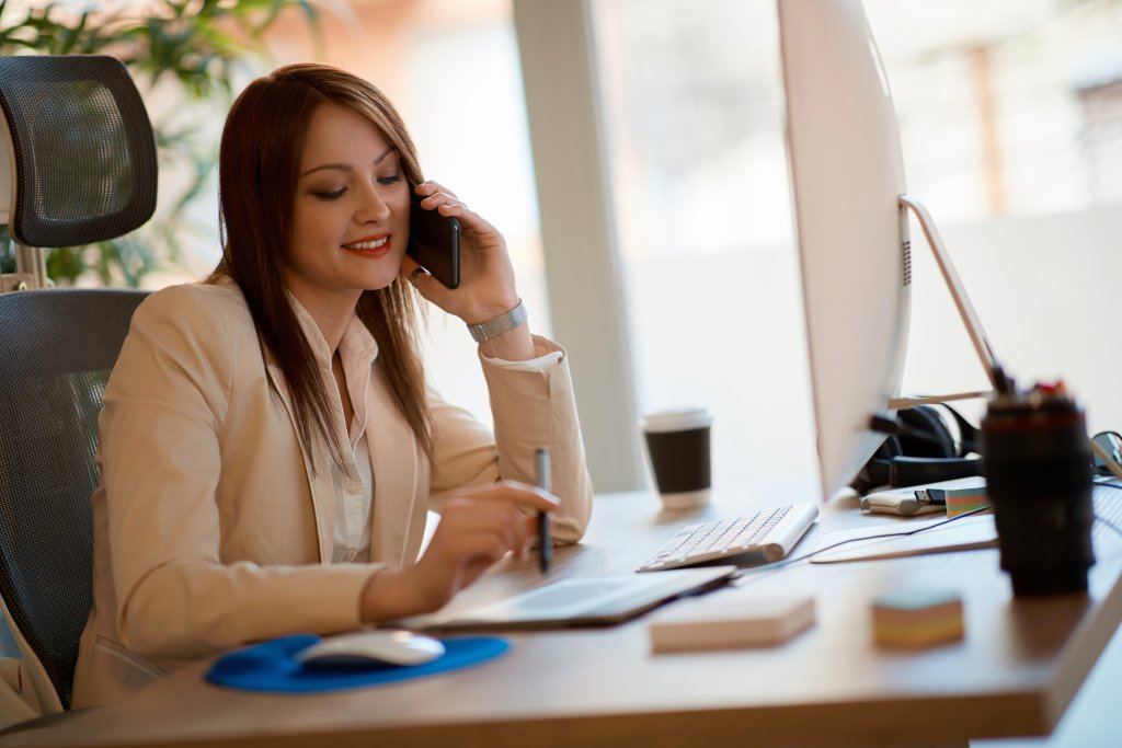 Eine l&auml;chelnde Frau in Gesch&auml;ftskleidung sitzt an ihrem Homeoffice-Schreibtisch, telefoniert und schreibt auf einem Notizblock. Vor ihr stehen ein Computer, eine Kaffeetasse und B&uuml;romaterialien - ein Beispiel daf&uuml;r, wie man bei COVID-19 richtig arbeitet.