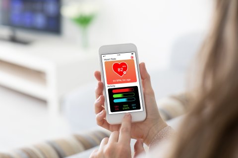 Eine Person h&auml;lt ein Smartphone in der Hand, auf dem eine Gesundheits-Apps-Benutzeroberfl&auml;che mit Herzfrequenz (82 BPM) und t&auml;glichen Aktivit&auml;tsstatistiken - Kalorien, Schritte und Entfernung - gut sichtbar angezeigt wird.