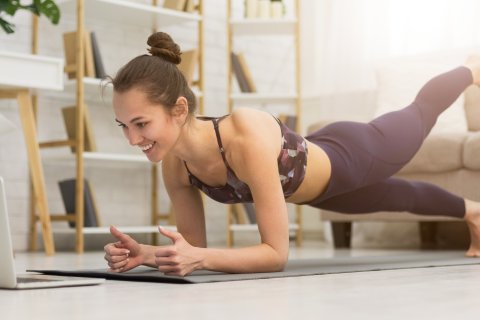 Eine Frau in Trainingskleidung macht eine Planke mit einem angehobenen Bein, w&auml;hrend sie in einem hellen Wohnzimmer l&auml;chelnd auf einen Laptop auf dem Boden schaut, was darauf schlie&szlig;en l&auml;sst, dass sie einen Online-Fitnesskurs besucht.