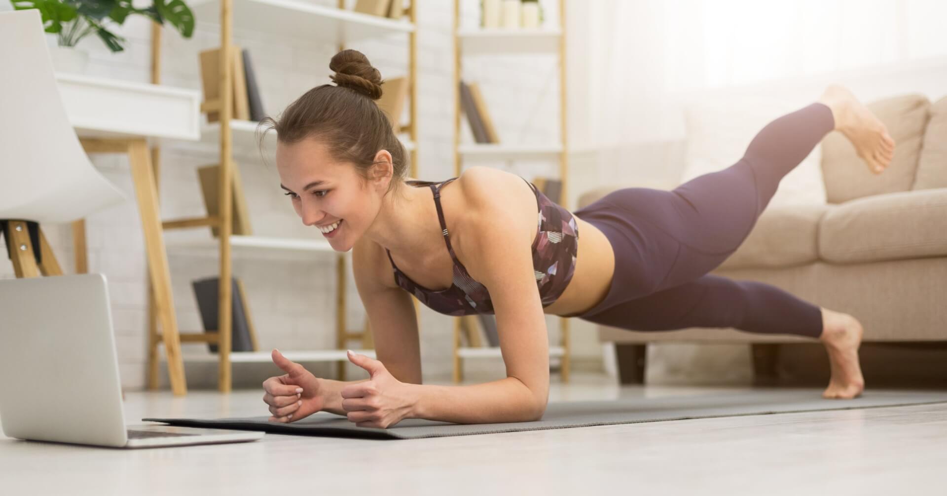 Eine Frau in Trainingskleidung macht eine Planke mit einem angehobenen Bein, w&auml;hrend sie in einem hellen Wohnzimmer l&auml;chelnd auf einen Laptop auf dem Boden schaut, was darauf schlie&szlig;en l&auml;sst, dass sie einen Online-Fitnesskurs besucht.