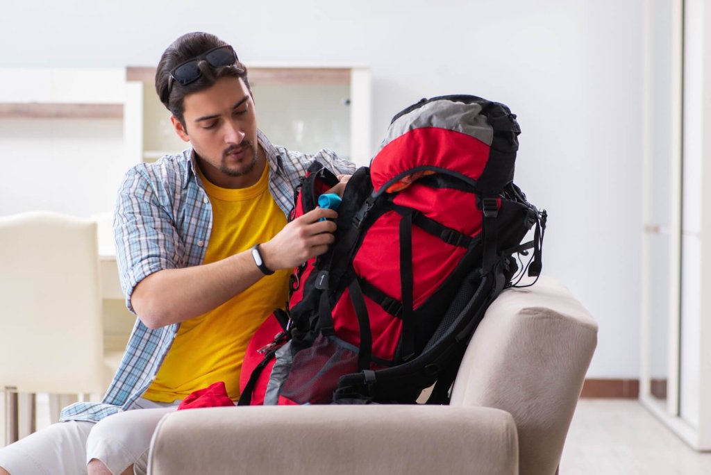 Ein Mann sitzt auf einer Couch, tr&auml;gt ein gelbes Hemd und eine Sonnenbrille auf dem Kopf und packt einen gro&szlig;en rot-schwarzen Rucksack in ein helles, modernes Wohnzimmer.