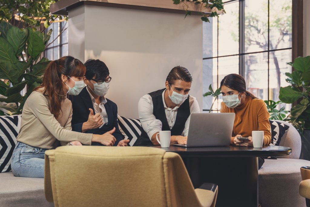 Vier Personen mit Gesichtsmasken sitzen um einen Tisch in einer B&uuml;rolounge, schauen auf einen Laptop-Bildschirm und diskutieren dar&uuml;ber, wie man sich an die Post-COVID-19-Normalit&auml;t anpassen kann, wobei im Hintergrund Kaffeetassen und Pflanzen zu sehen sind.