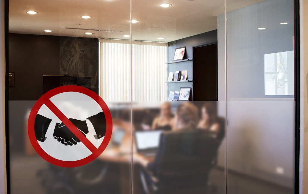 An einer gl&auml;sernen B&uuml;rowand h&auml;ngt ein Handschlagverbotsschild mit einem roten Kreis und einem Schr&auml;gstrich, das die COVID-19-Vorsichtsma&szlig;nahmen widerspiegelt. Hinter dem Glas sitzen mehrere Personen an einem Konferenztisch mit Laptops und passen sich der neuen Normalit&auml;t an.