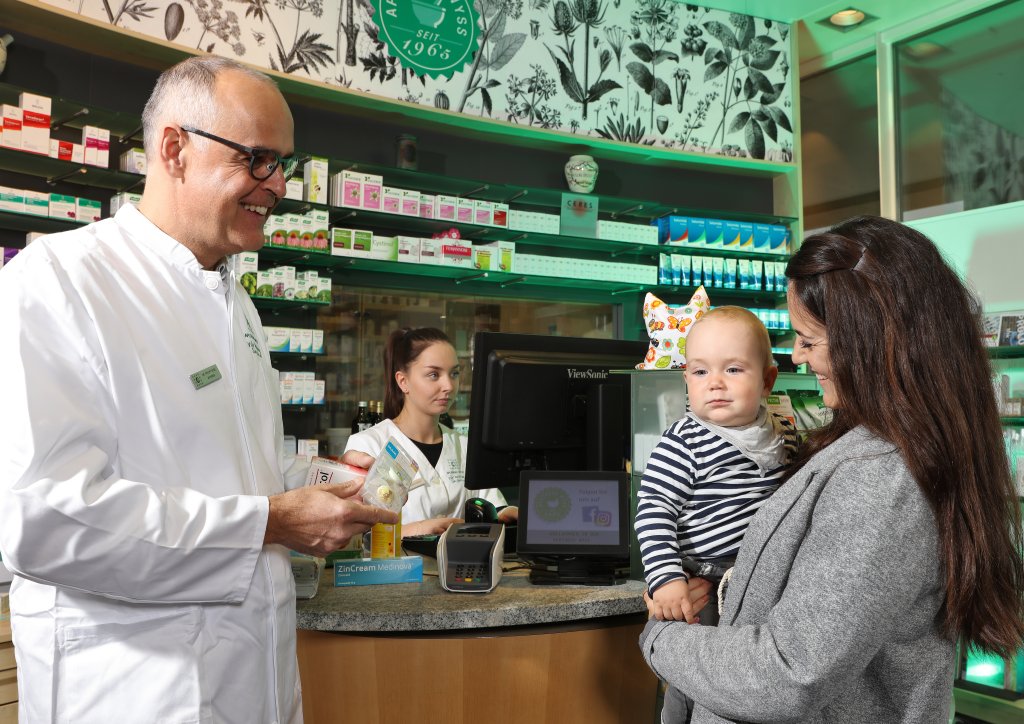 Ein Apotheker in einem wei&szlig;en Kittel l&auml;chelt und spricht mit einer Frau, die ein Baby in einer Apotheke h&auml;lt, w&auml;hrend ein anderer Mitarbeiter hinter dem Tresen am Computer arbeitet. Im Hintergrund sind Regale mit Medikamenten und Gesundheitsprodukten zu sehen.