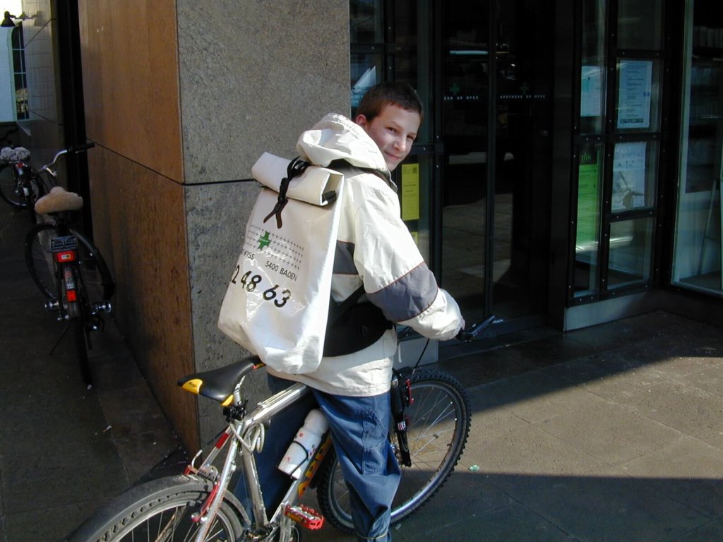 Ein Junge mit einer wei&szlig;-grauen Jacke und einem gro&szlig;en Lieferservice-Rucksack sitzt auf einem Fahrrad in der N&auml;he eines Hauseingangs, blickt &uuml;ber seine Schulter zur&uuml;ck und l&auml;chelt in die Kamera.