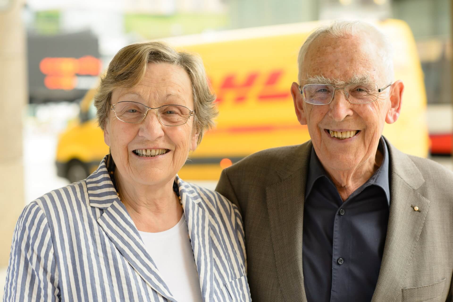 Eine &auml;ltere Frau und ein &auml;lterer Mann stehen im Freien dicht beieinander und l&auml;cheln in die Kamera. Im Hintergrund, in der N&auml;he der Stammapotheke, ist ein gelber DHL-Lieferwagen zu sehen.