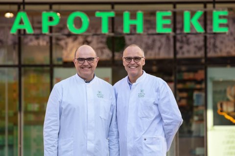 Zwei l&auml;chelnde M&auml;nner in wei&szlig;en Laborkitteln stehen vor einem Geb&auml;ude mit einem gro&szlig;en gr&uuml;nen Apothekenschild, das auf ihre Stammapotheke hinweist. Sie scheinen Apotheker oder Angestellte zu sein.