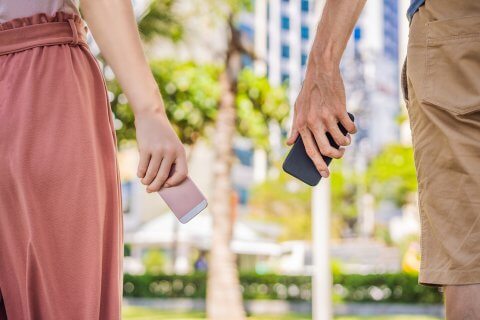 Zwei Personen stehen im Freien nebeneinander und halten jeweils ein Smartphone in der Hand - perfekt, um die SwissCovid App oder andere Tools zur Kontaktsuche zu nutzen. Die linke Person tr&auml;gt einen pinken Rock und h&auml;lt ein pinkes Telefon, die rechte eine hellbraune Shorts und ein schwarzes Telefon.