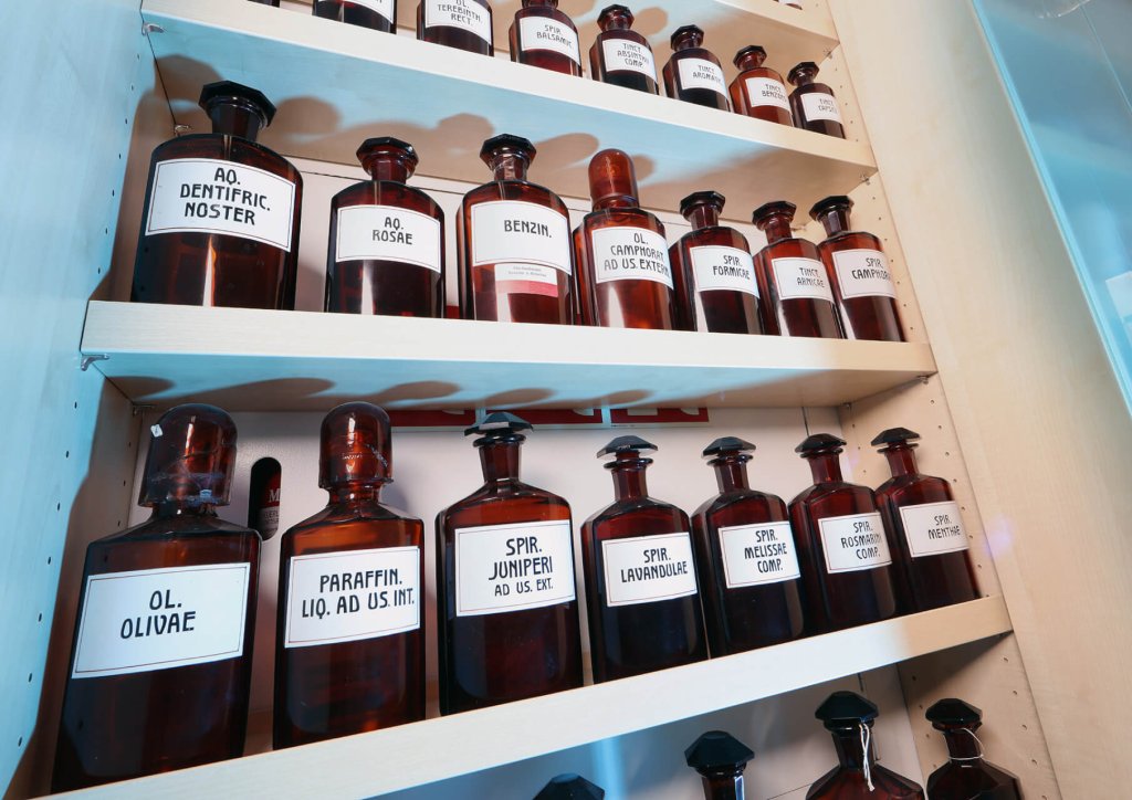 Die Regale sind gef&uuml;llt mit zahlreichen braunen Glasflaschen mit lateinischen Etiketten, die f&uuml;r Medikamente in einer altmodischen Apotheke oder einem Labor verwendet werden. Die Etiketten enthalten Namen wie OL. OLIVAE und SPIR. JUNIPERI, die eine korrekte Aufbewahrung widerspiegeln.