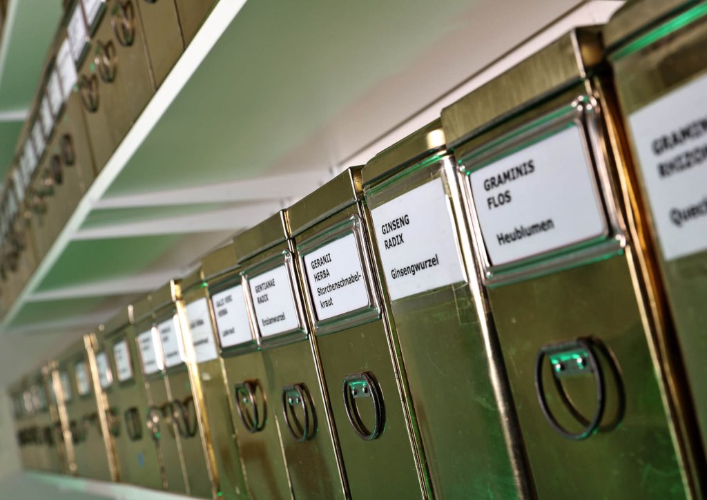 Metalldosen mit beschrifteten Etiketten, wie GINSENG RADIX und GRAMINIS FLOS, sind ordentlich auf wei&szlig;en Regalen angeordnet und demonstrieren die korrekte Lagerung von Medikamenten und Kr&auml;utern f&uuml;r eine optimale Organisation und Aufbewahrung.