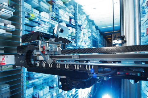 Ein Roboterarm entnimmt Medikamentenboxen in einem automatischen Apothekenlagersystem und sorgt f&uuml;r eine korrekte Lagerung. Sauber geordnete Reihen von Arzneimitteln werden durch helle, blau get&ouml;nte Beleuchtung f&uuml;r eine optimale Medikamentenaufbewahrung beleuchtet.