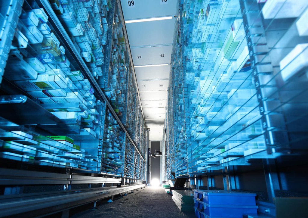 Ein schmaler Gang zwischen hohen, mit verschiedenen Medikamentenboxen gef&uuml;llten Regalen in einem hellen, modernen, mit blauem Licht beleuchteten Apothekenlagerraum sorgt f&uuml;r korrekte Lagerung und sichere Medikamentenaufbewahrung.