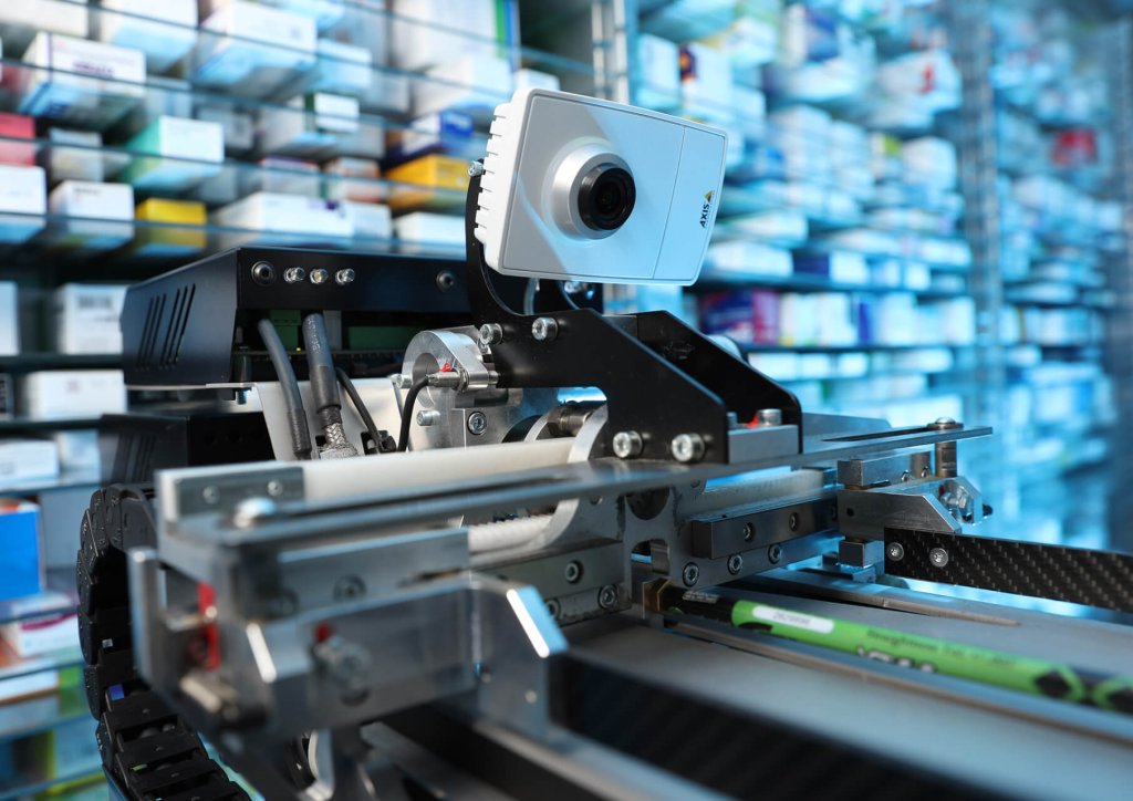 Nahaufnahme eines Roboterarms mit einer Kamera in einer Apotheke, umgeben von Regalen f&uuml;r Medikamentenaufbewahrung. Der unscharfe Hintergrund unterstreicht die Pr&auml;zision und fortschrittliche Technologie der Maschine f&uuml;r korrektes Lagern von Medikamenten.