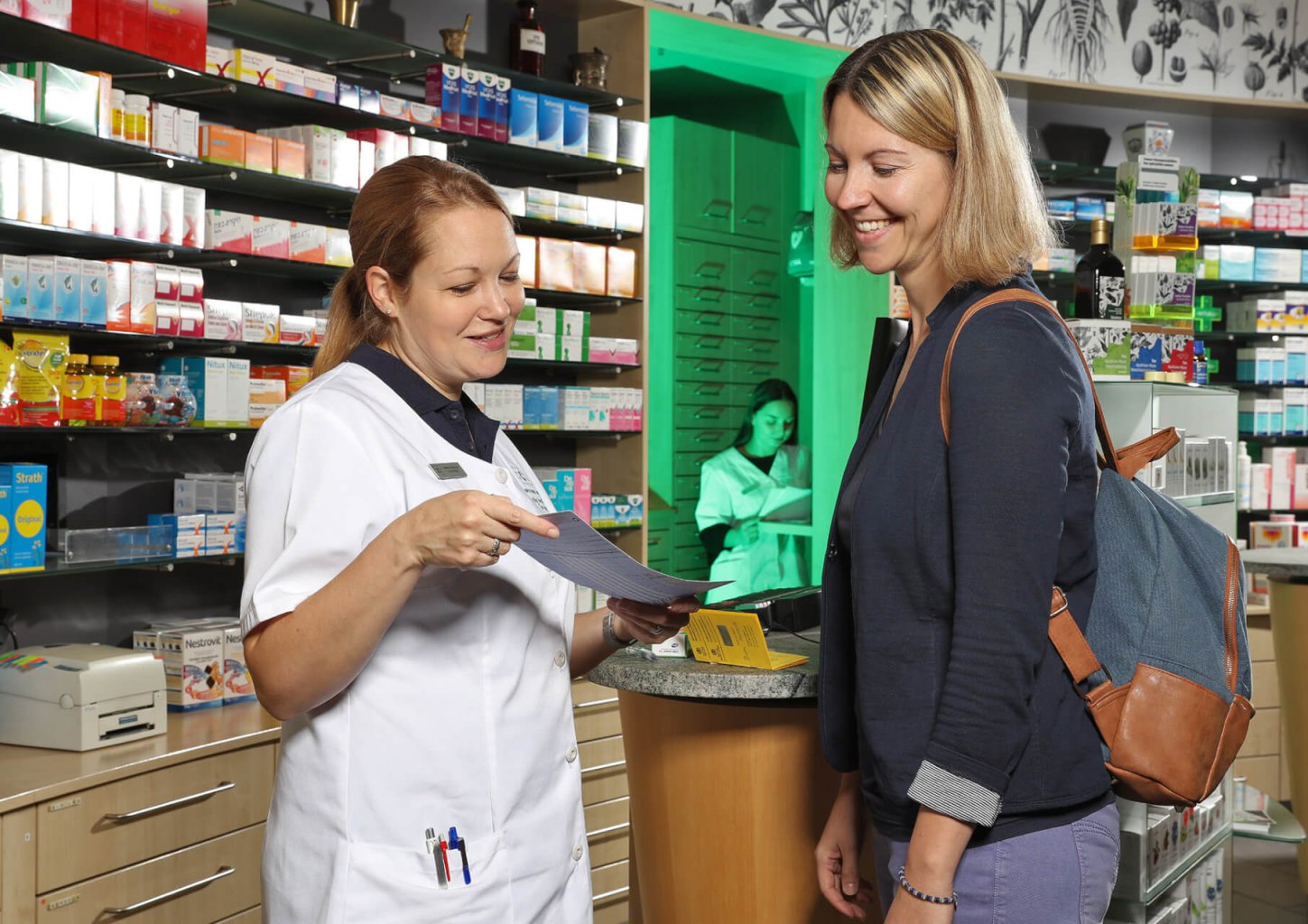Ein Apotheker im wei&szlig;en Kittel erkl&auml;rt einer l&auml;chelnden Kundin mit Rucksack in einer Apotheke Informationen aus einem Faltblatt &uuml;ber das neue Heilmittelgesetz, w&auml;hrend im Hintergrund Regale mit Arzneimitteln und eine weitere Mitarbeiterin besch&auml;ftigt sind.