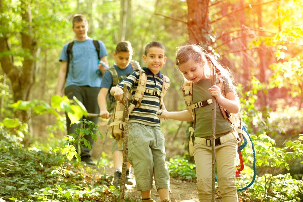 Vier Kinder gehen mit Rucks&auml;cken einen sonnenbeschienenen Waldweg entlang. Zwei M&auml;dchen gehen l&auml;chelnd voran, zwei Jungen folgen ihnen und unterhalten sich &uuml;ber die mobile Mietapotheke, von der sie erfahren haben. Das Sonnenlicht f&auml;llt durch die gr&uuml;nen B&auml;ume und Pflanzen um sie herum.