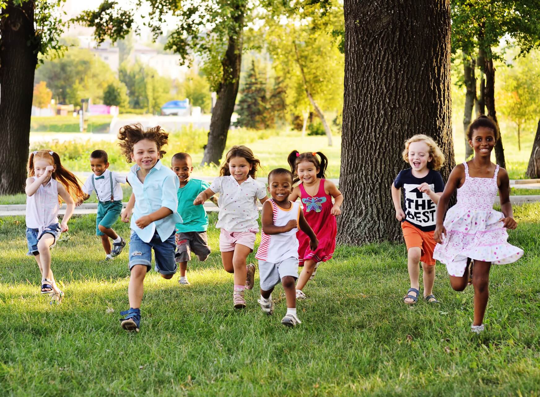 Eine Gruppe l&auml;chelnder Kinder unterschiedlicher Herkunft rennt an einem sonnigen Tag gemeinsam &uuml;ber eine Parkwiese, w&auml;hrend im Hintergrund zwischen B&auml;umen und Gr&uuml;npflanzen eine mobile Apotheke aufgebaut ist.