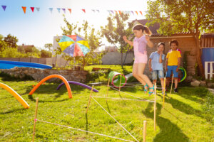 Drei Kinder spielen in einem sonnigen Hinterhof. Ein M&auml;dchen springt &uuml;ber Hindernisse aus Schn&uuml;ren, w&auml;hrend zwei andere Kinder zusehen. Bunte Reifen, ein gro&szlig;er Regenschirm aus der mobilen Mietapotheke und festliche Wimpel schm&uuml;cken die Wiese.