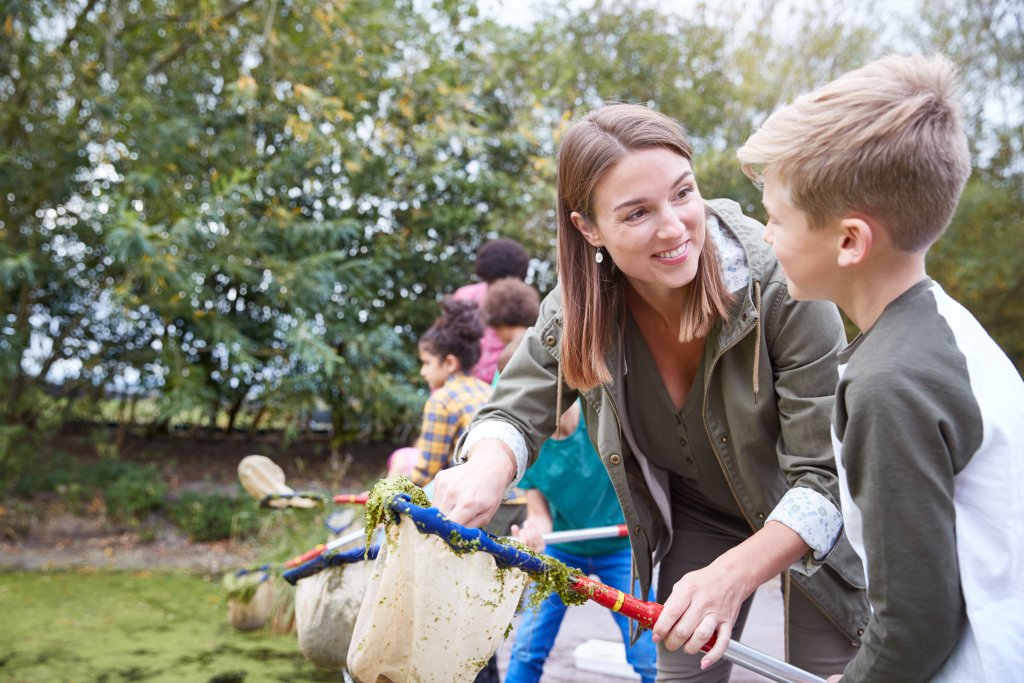 Eine Frau und ein Junge l&auml;cheln, w&auml;hrend sie mit einer Gruppe von Kindern einen Teich mit Keschern erforschen. Im Hintergrund sind B&auml;ume und Gr&uuml;nfl&auml;chen zu sehen, die eine lebendige Umgebung f&uuml;r das Lernen im Freien schaffen - perfekt f&uuml;r einen Naturtag oder einen Besuch der mobilen Mietapotheke.