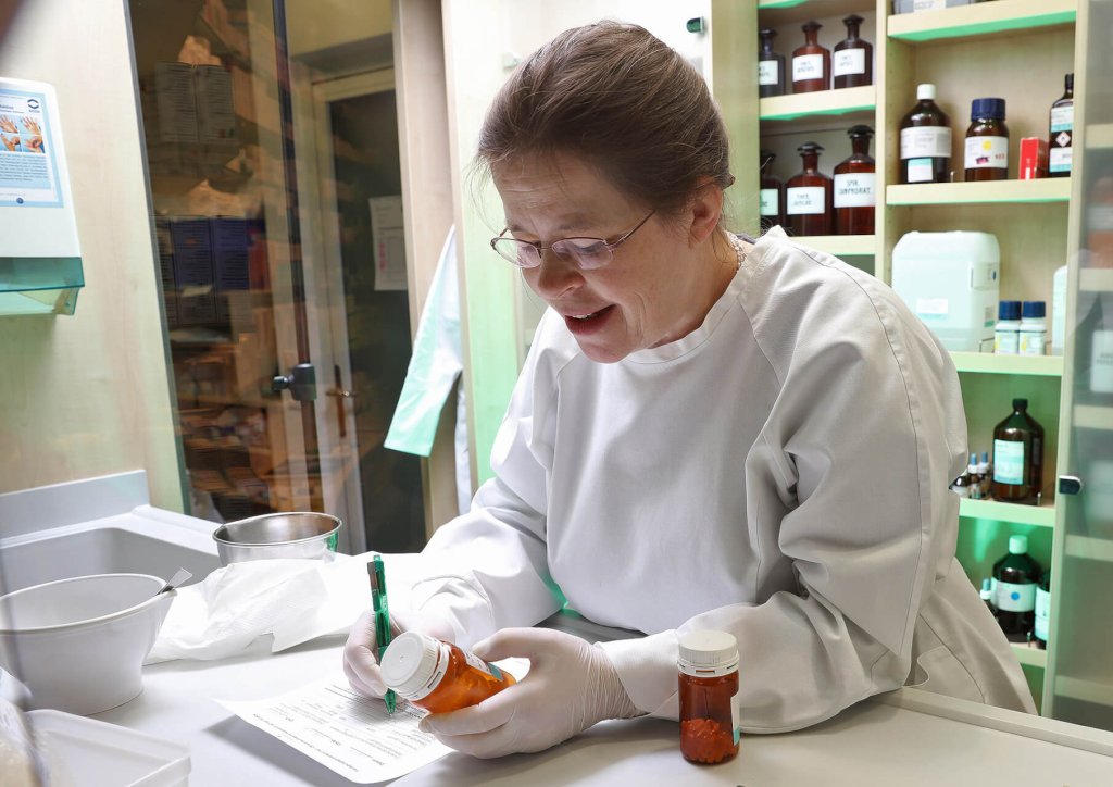 Eine Apothekerin im wei&szlig;en Kittel und mit Handschuhen etikettiert eine Rezeptflasche an einem Apothekentresen, umgeben von Arzneimittelflaschen, Papierkram und QMS-Dokumenten. Sie l&auml;chelt und konzentriert sich auf ihre Aufgabe.