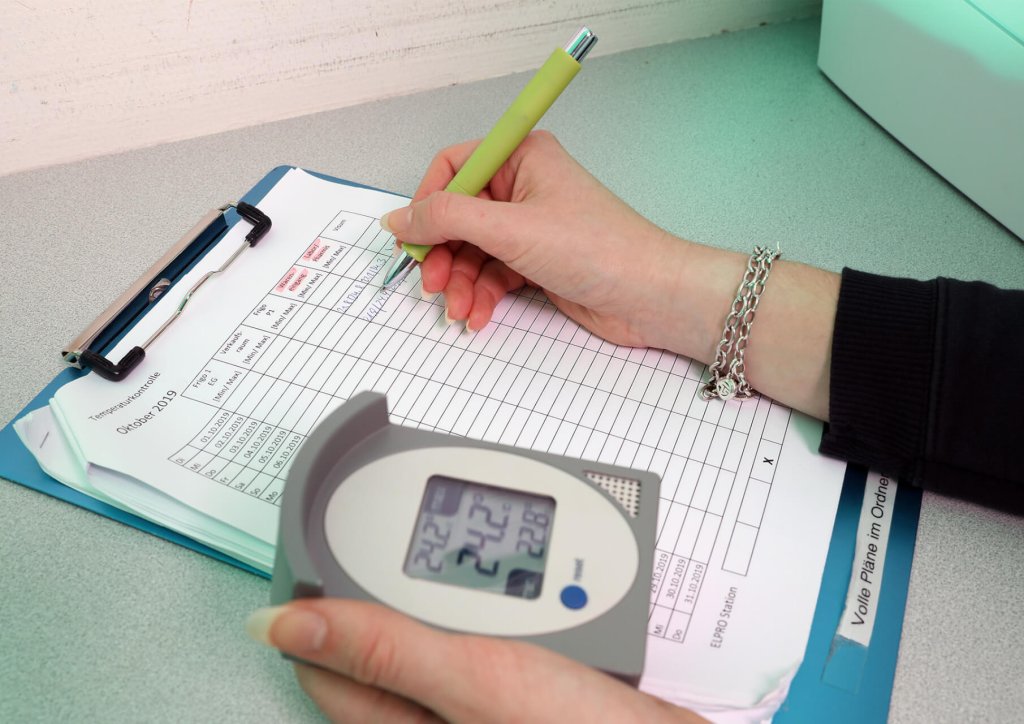 Eine Person aktualisiert ein Temperaturprotokoll auf einem Klemmbrett mit einem Stift, w&auml;hrend sie ein digitales Thermometer in der Hand h&auml;lt, ein wesentlicher Schritt in QMS- und Qualit&auml;tsmanagementverfahren.