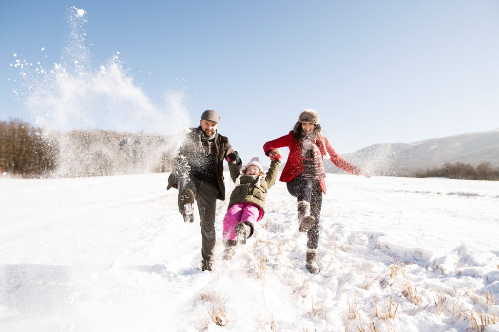 Eine dreik&ouml;pfige Familie genie&szlig;t einen nat&uuml;rlichen Wintertag und spielt im Schnee. Die Eltern schwingen ihr Kind zwischen sich, alle l&auml;cheln in warmer Kleidung, w&auml;hrend der Schnee herumfliegt. H&uuml;gel und B&auml;ume vervollst&auml;ndigen die gesunde Outdoor-Szene.