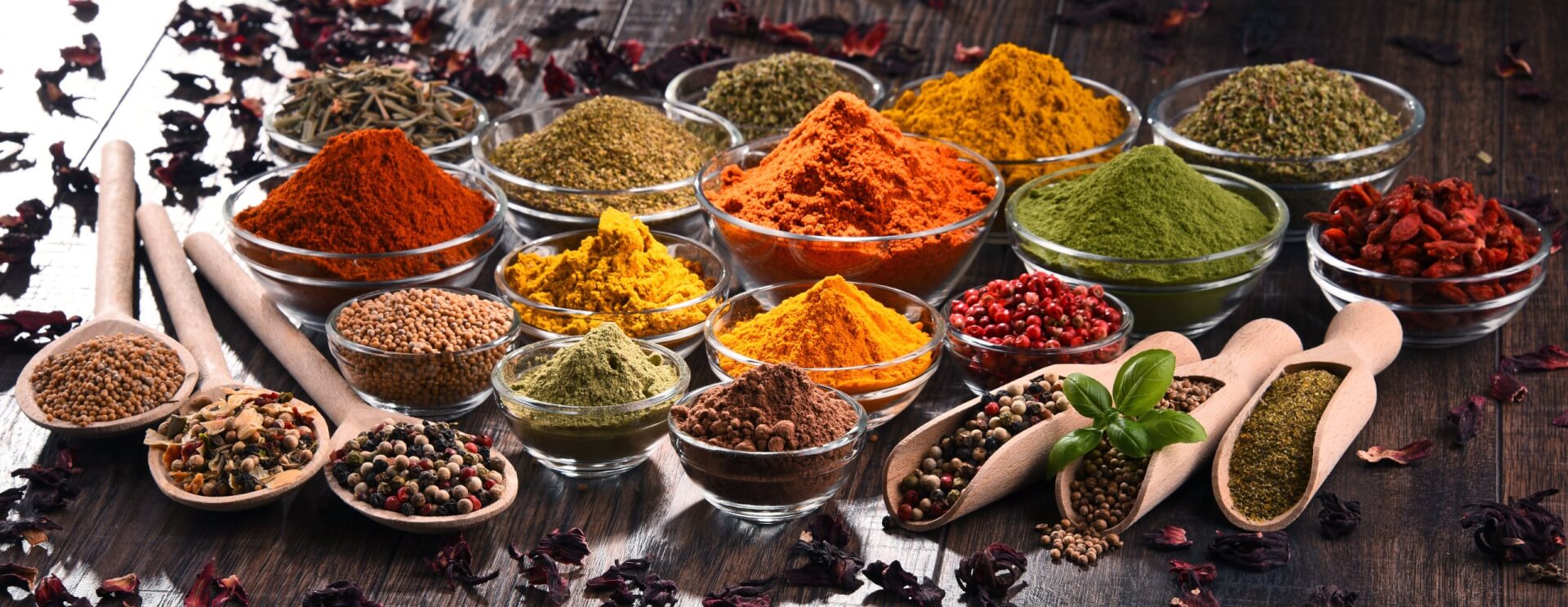 Eine Vielzahl bunter Gew&uuml;rze und Kr&auml;uter, die als Heilmittel gesch&auml;tzt werden, sind in kleinen Glassch&auml;lchen und Holzl&ouml;ffeln auf einer rustikalen Holzfl&auml;che ausgestellt, darunter Pulver und ganze Samen in Rot-, Gelb-, Gr&uuml;n- und Braunt&ouml;nen.