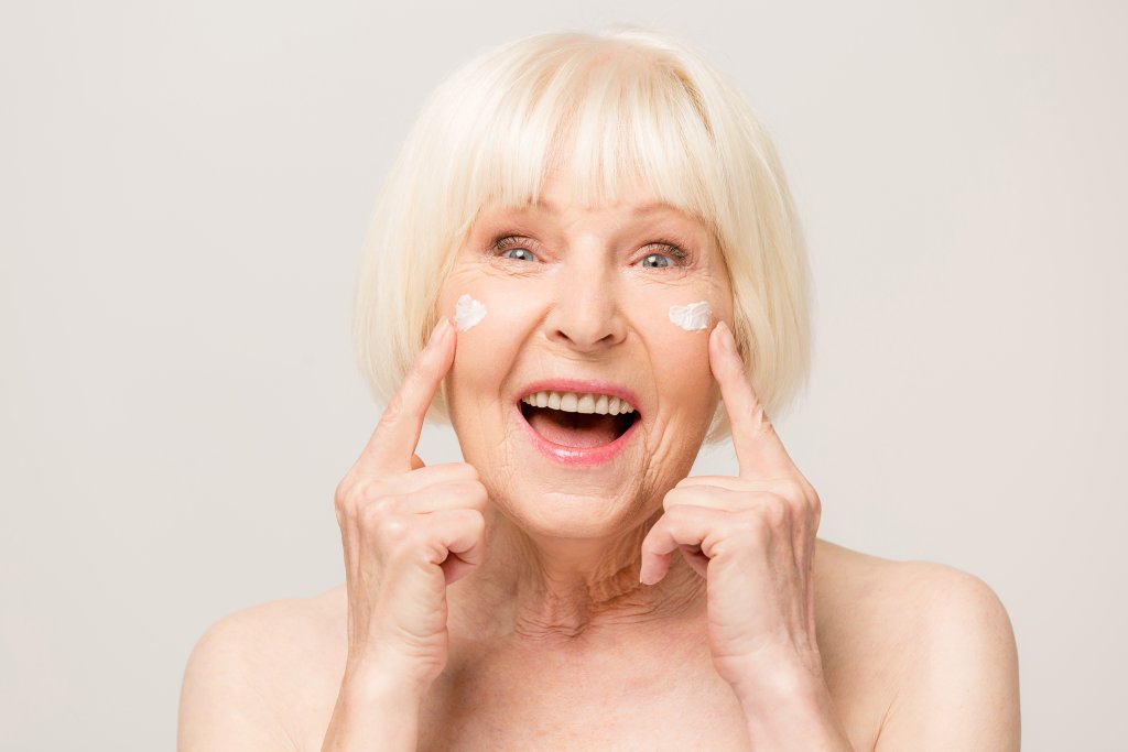 Eine &auml;ltere Frau mit kurzen wei&szlig;en Haaren l&auml;chelt und zeigt auf eine Creme, die unter beiden Augen aufgetragen wird, um zu zeigen, wie sie trockene Haut behandeln und ihre Hautpflege-Routine genie&szlig;en kann, vor einem schlichten hellen Hintergrund.