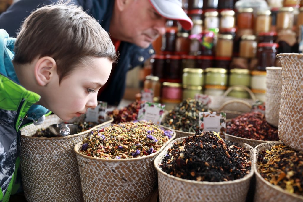 Ein kleiner Junge riecht auf einem Markt intensiv an einem Korb mit bunten Gew&uuml;rzen, w&auml;hrend ein erwachsener Mann mit einer roten M&uuml;tze zuschaut. Im Hintergrund sind Gl&auml;ser mit Konserven, weitere K&ouml;rbe mit Gew&uuml;rzen und traditionelle Heilmittel zu sehen.