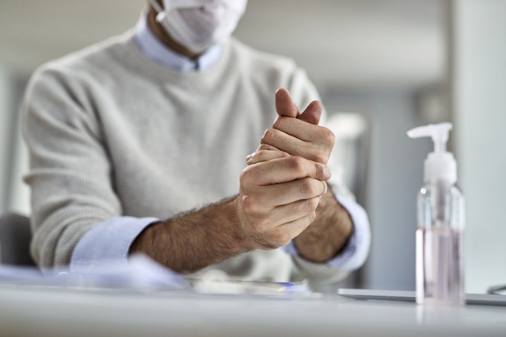 Eine Person, die eine Gesichtsmaske und einen leichten Pullover tr&auml;gt, reibt sich die H&auml;nde neben einer Flasche mit Handdesinfektionsmittel auf einem Tisch, um auf gute Hygienepraktiken und das Bewusstsein f&uuml;r Grippeimpfung in Innenr&auml;umen hinzuweisen.