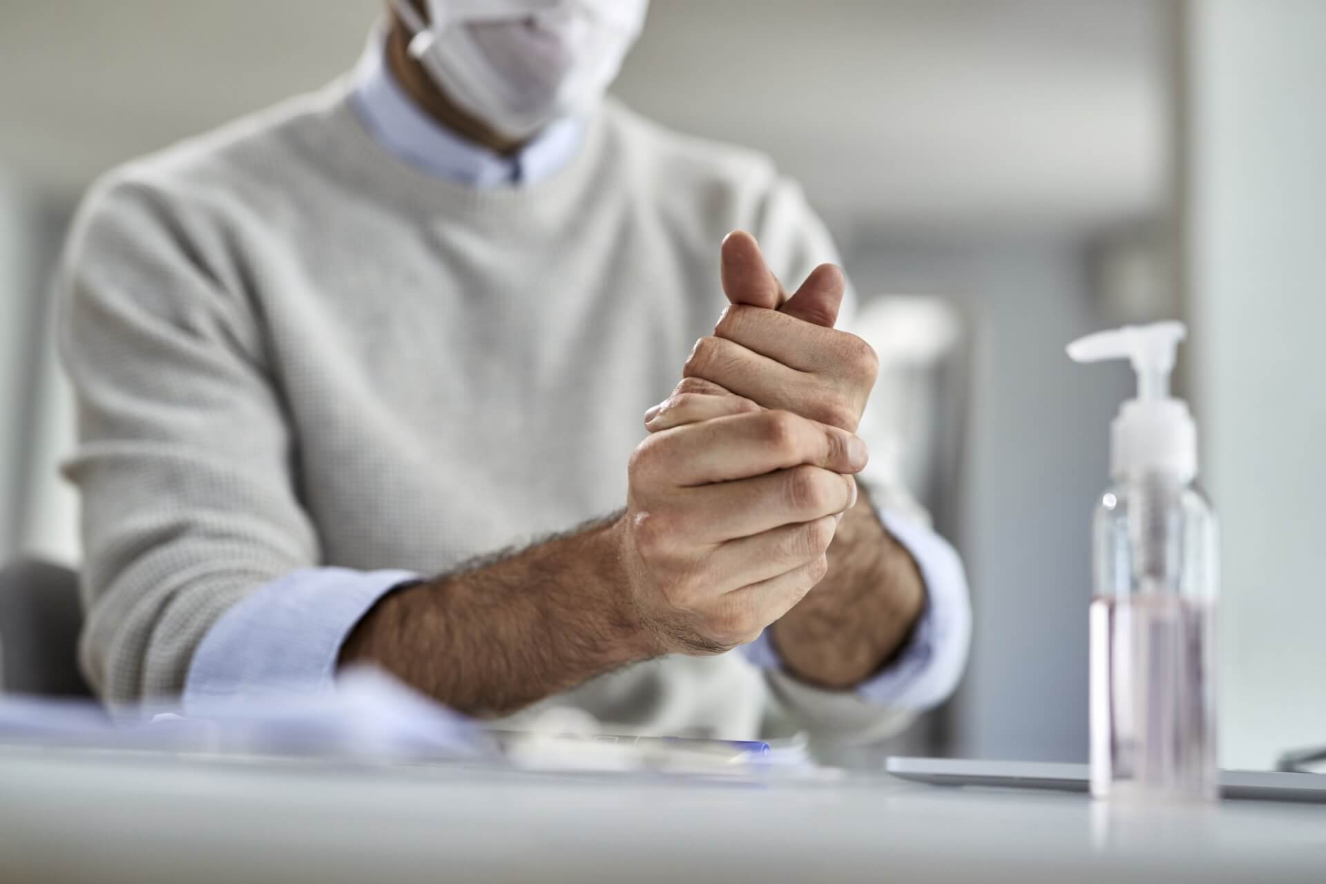 Eine Person, die eine Gesichtsmaske und einen leichten Pullover tr&auml;gt, reibt sich die H&auml;nde neben einer Flasche mit Handdesinfektionsmittel auf einem Tisch, um auf gute Hygienepraktiken und das Bewusstsein f&uuml;r Grippeimpfung in Innenr&auml;umen hinzuweisen.