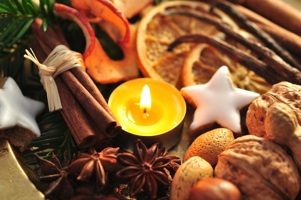 Ein brennendes Teelicht, umgeben von Gew&uuml;rzen wie Zimtstangen, Sternanis und Vanilleschoten sowie Waln&uuml;ssen, getrockneten Orangenscheiben, Mandeln und sternf&ouml;rmigen Eispl&auml;tzchen, schafft eine festliche und gem&uuml;tliche Atmosph&auml;re.