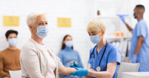 Eine medizinische Fachkraft in blauem Kittel verabreicht einer &auml;lteren Frau, die eine Maske tr&auml;gt, eine Grippeimpfung. Andere Maskierte sitzen im Hintergrund, und eine weitere medizinische Fachkraft steht in der N&auml;he.