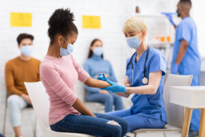 Eine Krankenschwester in blauem Kittel bereitet den Arm einer jungen Frau f&uuml;r eine Grippeimpfung vor. Beide tragen Masken. Im Hintergrund warten drei weitere maskierte Personen in einem hell erleuchteten Raum auf ihren Einsatz.