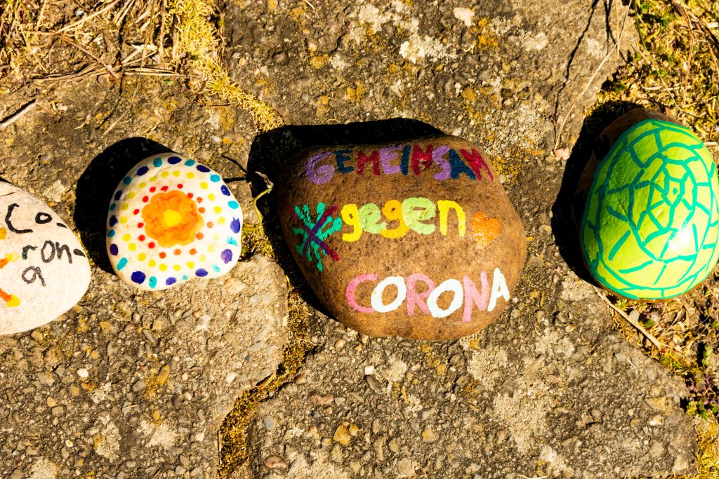 Vier bemalte Felsen auf einem Gehweg im Freien; ein Felsen zeigt die bunten deutschen Worte "gegen CORONA" mit hellen Mustern, die auf COVID-19 verweisen, w&auml;hrend andere Punkte, Muster und ein gr&uuml;nes geometrisches Muster aufweisen. Das Sonnenlicht wirft Schatten auf die Szene.