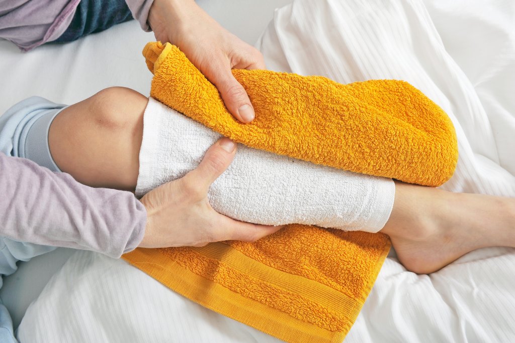Eine Person wickelt ein gelbes Handtuch um ein bandagiertes Knie, w&auml;hrend sie auf einem Bett sitzt, was darauf hindeutet, dass sie eine Verletzung oder ein geschwollenes Knie mit Wickel oder Umschl&auml;gen versorgt.