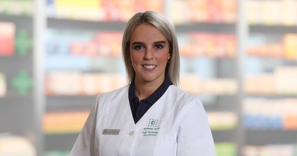 Alice Troisi, die einen wei&szlig;en Laborkittel und ein Namensschild tr&auml;gt, steht l&auml;chelnd vor unscharfen Regalen, die auf eine Apotheke oder einen Gesundheitsbereich hindeuten.