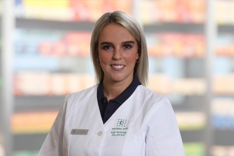 Alice Troisi, die einen wei&szlig;en Laborkittel und ein Namensschild tr&auml;gt, steht l&auml;chelnd vor unscharfen Regalen, die auf eine Apotheke oder einen Gesundheitsbereich hindeuten.