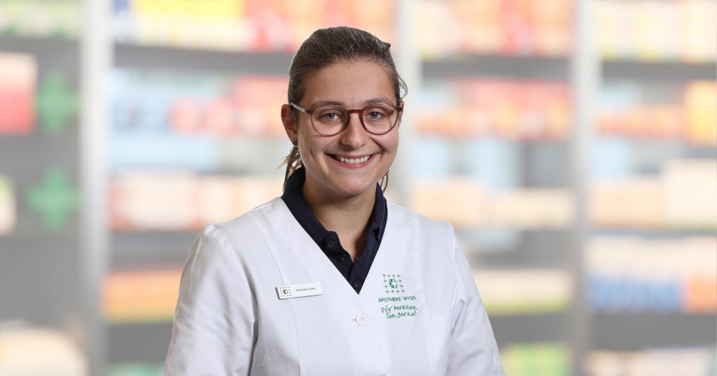 Eine l&auml;chelnde Frau, Anastasia Fonte, mit Brille und wei&szlig;em Laborkittel steht vor verschwommenen Regalen, wahrscheinlich in einer Apotheke oder einem Gesch&auml;ft. Sie hat ihr dunkles Haar zur&uuml;ckgebunden und tr&auml;gt ein Namensschild an ihrem Kittel.