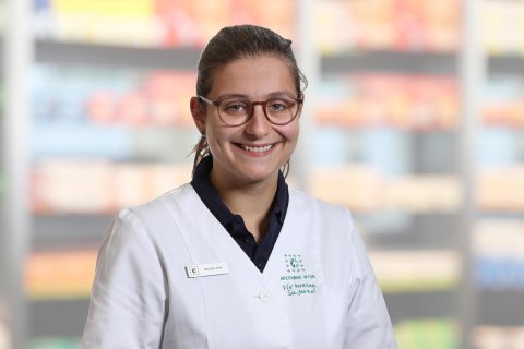 Eine l&auml;chelnde Frau, Anastasia Fonte, mit Brille und wei&szlig;em Laborkittel steht vor verschwommenen Regalen, wahrscheinlich in einer Apotheke oder einem Gesch&auml;ft. Sie hat ihr dunkles Haar zur&uuml;ckgebunden und tr&auml;gt ein Namensschild an ihrem Kittel.