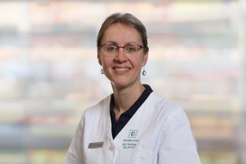 Anita Zimmermann, die eine Brille und einen wei&szlig;en Laborkittel mit einem Namensschild tr&auml;gt, l&auml;chelt in die Kamera. Der Hintergrund ist unscharf und farbenfroh, was auf eine Innenraumumgebung hindeutet.