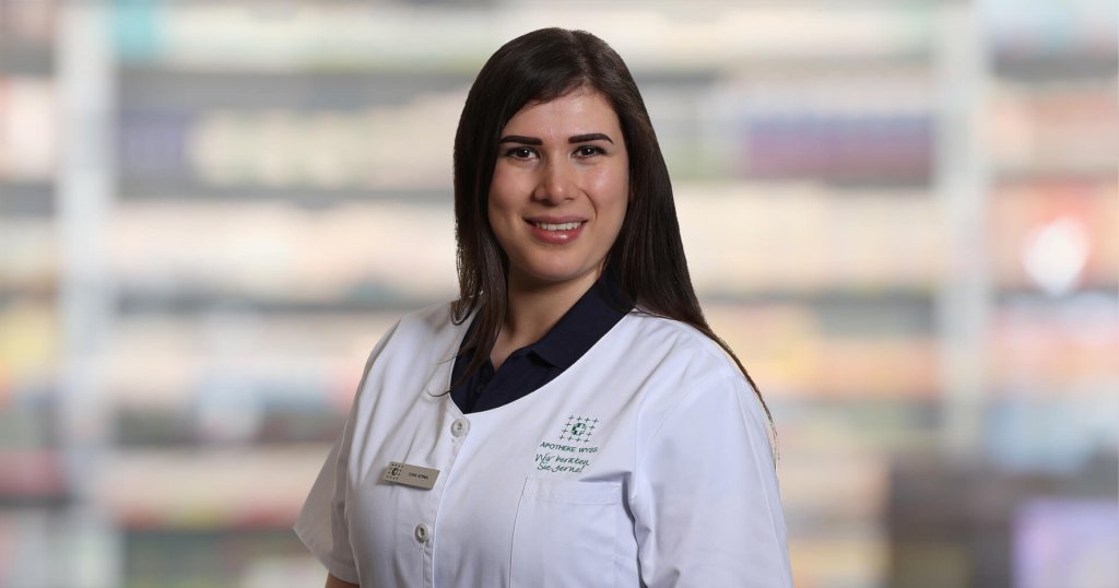 Bermal Demir, eine Frau mit langen dunklen Haaren, die einen wei&szlig;en Laborkittel und ein Namensschild tr&auml;gt, steht l&auml;chelnd vor verschwommenen Regalen, wahrscheinlich in einer Apotheke oder einem medizinischen Bereich.