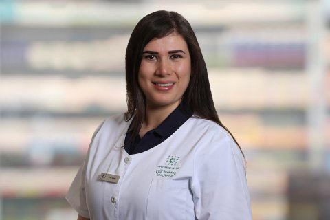 Bermal Demir, eine Frau mit langen dunklen Haaren, die einen wei&szlig;en Laborkittel und ein Namensschild tr&auml;gt, steht l&auml;chelnd vor verschwommenen Regalen, wahrscheinlich in einer Apotheke oder einem medizinischen Bereich.