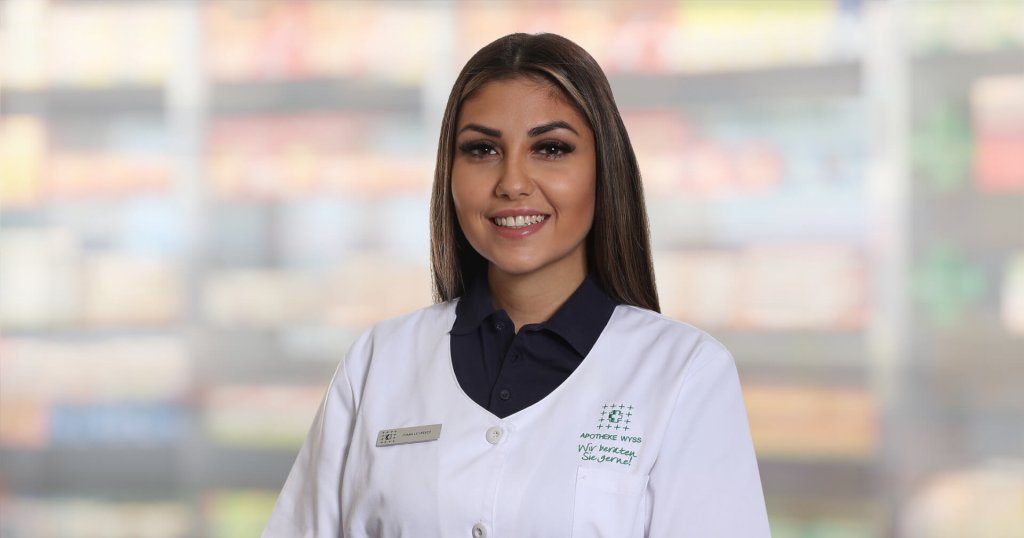 Joana Lourenco, die einen wei&szlig;en Apothekenkittel und ein dunkles Hemd tr&auml;gt, steht l&auml;chelnd vor verschwommenen Regalen. Sie hat langes braunes Haar, ein Namensschild und scheint sich in einer Apotheke oder einem Gesch&auml;ft zu befinden.