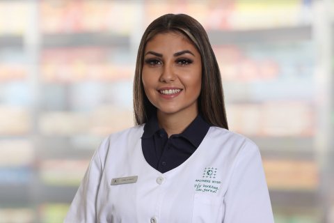 Joana Lourenco, die einen wei&szlig;en Apothekenkittel und ein dunkles Hemd tr&auml;gt, steht l&auml;chelnd vor verschwommenen Regalen. Sie hat langes braunes Haar, ein Namensschild und scheint sich in einer Apotheke oder einem Gesch&auml;ft zu befinden.