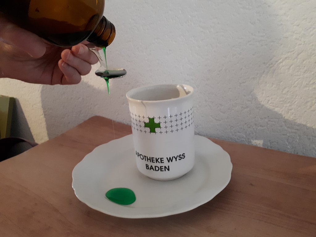 Eine Hand gie&szlig;t zuckerfreien Sirup aus einer braunen Flasche auf einen L&ouml;ffel &uuml;ber einen wei&szlig;en Becher mit der Aufschrift "APOTHEKE WYSS BADEN". Etwas gr&uuml;ne Fl&uuml;ssigkeit schwappt auf einen wei&szlig;en Teller unterhalb des Bechers, der auf einer h&ouml;lzernen Oberfl&auml;che steht.