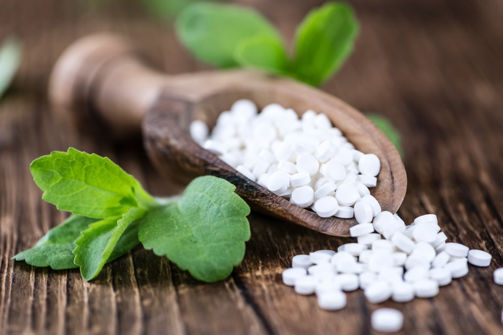 Ein Holzl&ouml;ffel h&auml;lt einen Stapel kleiner wei&szlig;er zuckerfreier Tabletten, von denen einige auf eine rustikale Holzfl&auml;che gestreut sind. In der N&auml;he liegen gr&uuml;ne Steviabl&auml;tter, die darauf hindeuten, dass die Tabletten ein S&uuml;&szlig;ungsmittel oder eine Alternative zu Sirup sind.